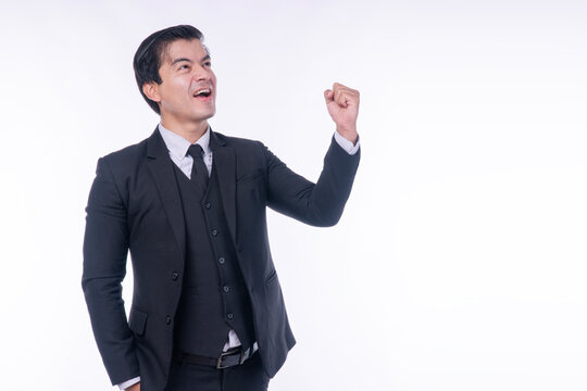 Excited handsome businessman in dark suit raising fist in victory and looking up at copy space on white background. Successful executive celebrating goal achievement or winning concept.