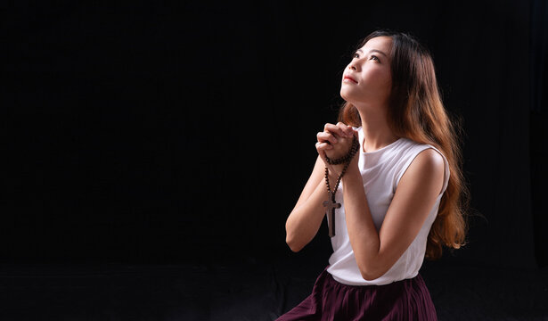 Young Asian woman in white top kneeling and praying with hands clasped holding rosary cross on dark black background with copy space. Spiritual female seeking hope and god blessing.