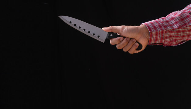 Close up of man hand in red plaid shirt holding sharp kitchen knife on dark black background with copy space. Dangerous criminal threat, horror murder or aggressive violence concept.