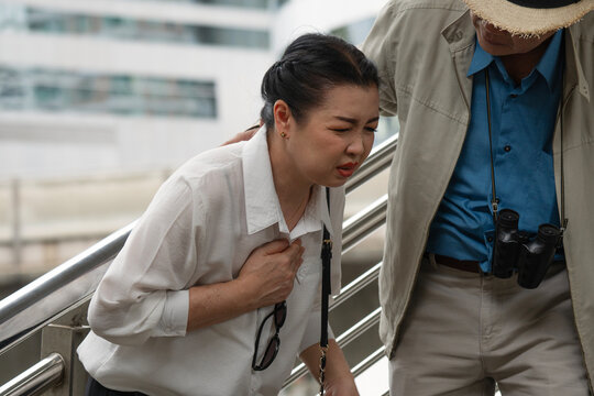Senior Asian woman having heart attack or chest pain walking down stairs in city. Elderly man helping his wife with sudden illness. Healthcare, medical and insurance concept.