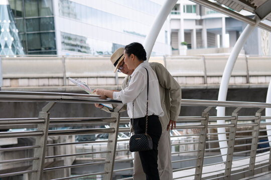 Senior Asian couple traveling in modern city standing on overpass looking at paper map to find direction. Elderly husband and wife tourists enjoying retirement vacation trip together in urban city.
