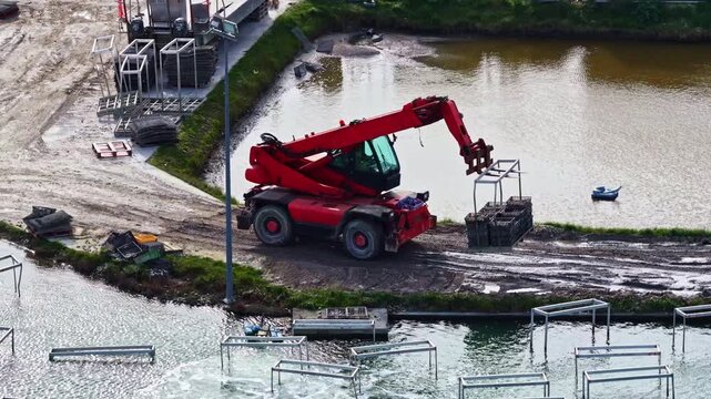 Aerial view of a red telehandler lifting a crate near a pond with metal frames at a construction site in France.