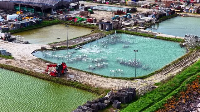 Aerial view of an oyster farm featuring aquaculture ponds, submerged metal racks, and a red telehandler on the embankment in France.