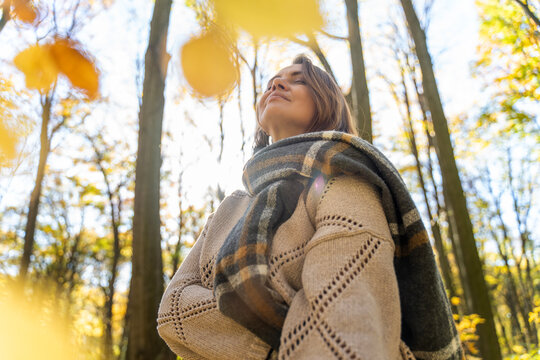 Mature Caucasian woman in warm sweater and scarf enjoying fresh air in autumn forest. Mental health and healthy aging concept