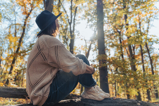 Beautiful thoughtful mature woman in fashion hat and cozy clothing looking away, sitting on nature during autumn weekend.