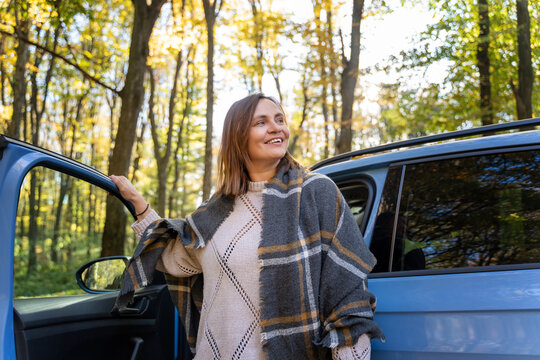 Cheerful mature woman wrapped in plaid blanket taking a break near vehicle, enjoying autumn sunshine. Healthy aging concept