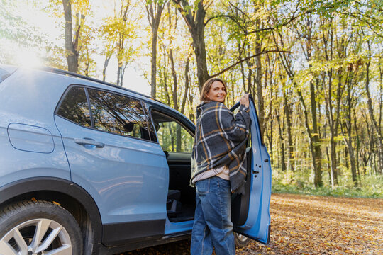 Cheerful mature woman wrapped in plaid blanket taking a break near vehicle, enjoying autumn sunshine. Healthy aging concept