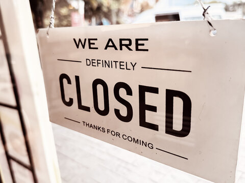 Close-up view of a white rectangular sign reading "WE ARE DEFINITELY CLOSED - THANKS FOR COMING" hanging on a glass door of a shop or restaurant.