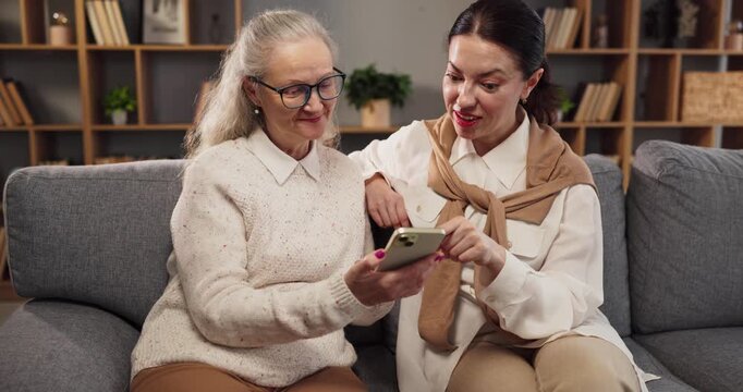 Senior woman and caregiver learning smartphone technology. At home the younger woman guides the senior through apps, building confidence and connection. Clear concept of digital caregiving.