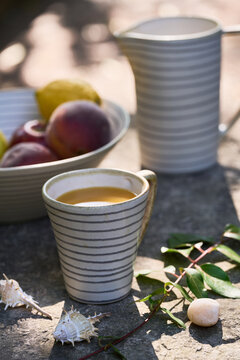 Close vertical product composition featuring a striped ceramic cup in the foreground, with a matching pitcher and bowl of fresh fruit softly blurred behind.