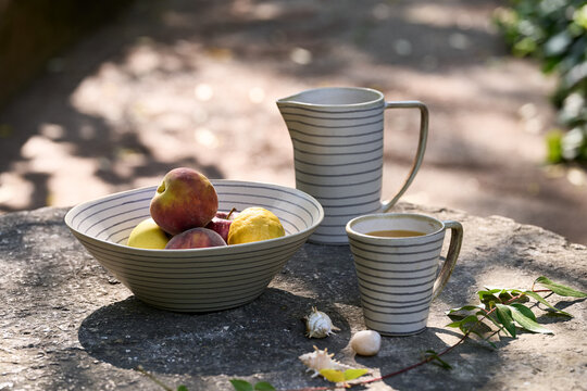 Rustic striped ceramic bowl, pitcher, cup and serving dish arranged with whole and sliced peaches, apples, lemon and orange juice on a stone table in dappled garden sunlight.