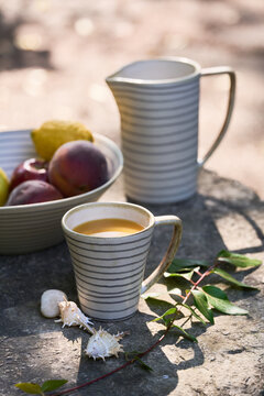 Close vertical product composition featuring a striped ceramic cup in the foreground, with a matching pitcher and bowl of fresh fruit softly blurred behind.