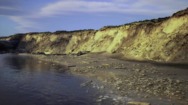 rugged cliff shoreline during ebb tide with warm side light carving deep shadows into stratified sandstone, exposed rock pools and wet sand reflecting sky,
