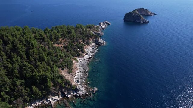 Aerial drone view of a crystal clear turquoise bay on Thassos Island, Greece, with swimmers, paddleboards and a sandy beach surrounded by rocky coastline on a sunny summer day.