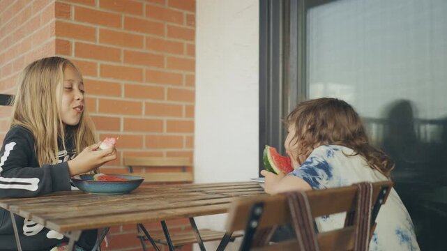 Two siblings laughing and eating watermelon together on a balcony