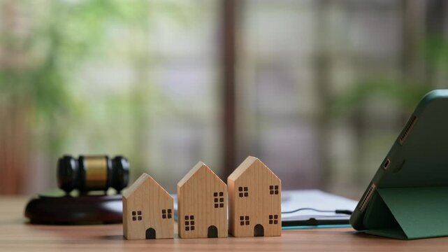 Three wooden house models stand beside legal judge gavel near tablet computer screen showing video playback interface inside bright office space representing real estate law litigation property.