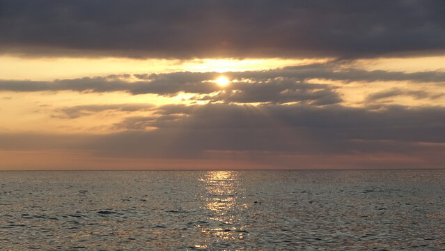 Bright Sun Peeking Through Cloud Layer with Golden Water Reflection.
A dramatic sunrise showing the sun breaking through thick cloud cover and creating a shimmering light path on the sea.