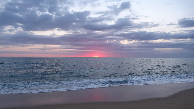 Vibrant Red Dawn Glow on the Sea Horizon.
A dramatic red dawn glow lighting up the cloudy sky right before the sun peeks above the ocean horizon on a quiet sand beach.