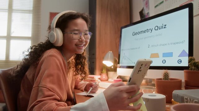 Asian teen girl wearing headphones studying geometry at desk, picking up smartphone to interact with educational app. Footage is ideal for e-learning platforms and remote education marketing