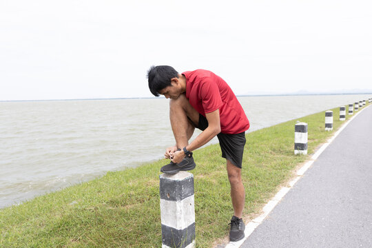 Asian male athlete tying running shoe while preparing for outdoor exercise, representing fitness, healthy lifestyle, training routine, and running preparation.