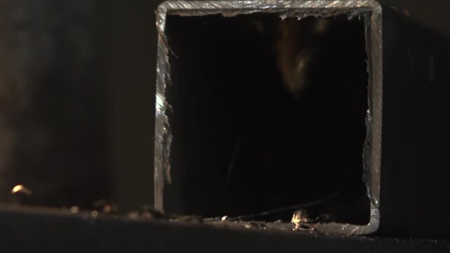 Close-up view of square hollow steel tube with freshly cut jagged rim, burrs, and curled metal shavings on workshop surface under low-key lighting, with metal, steel, tube, square detail