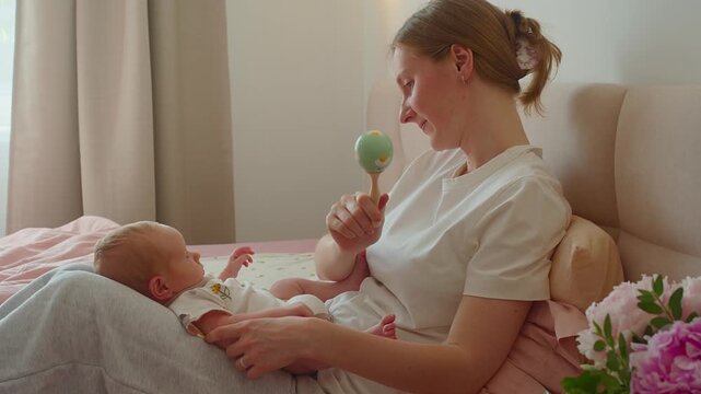 Mother moves rattle from side to side before baby on bed. Moving toy encourages visual tracking, focused attention, curiosity, sensory learning, and playful communication with parent.