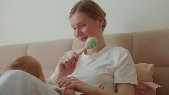 Mother shakes rattle from side to side before newborn during playful interaction. Rhythmic toy movement promotes tracking, attention, sensory stimulation, curiosity, and lively engagement.