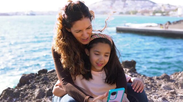 Happy mother and little daughter using mobile phone on the beach during summer vacation - Family lifestyle, bonding and love concept	