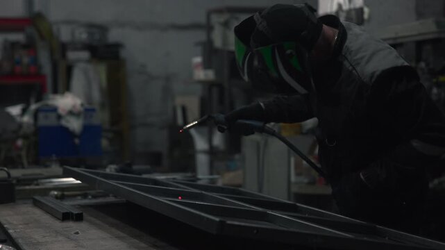 Welder positioning torch over steel beams on concrete workbench inside dim industrial workshop with protective gear, orange embers, and muted smoky atmosphere, with welder, welding, torch, metal