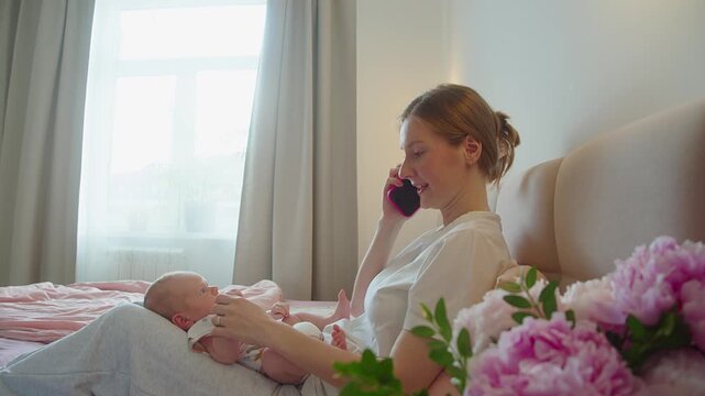 Mother talks on smartphone while infant rests against her body on bed. Close contact combines mobile communication, caregiving, comfort, security, maternal presence, and everyday family routine.