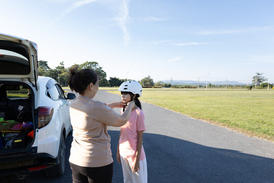 Asian mother adjusts a safety helmet for her young daughter during outdoor activity in a park, showing child safety, parental care, and family lifestyle.
