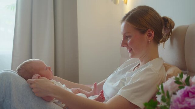 Mother talks to infant while gently holding baby hands on bed. Quiet speech and playful contact create emotional bonding, attentive communication, trust, comfort, and nurturing family care.