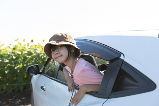 Asian girl leaning out from a car window during an outdoor trip, showing childhood curiosity, travel lifestyle, and relaxed family journey.