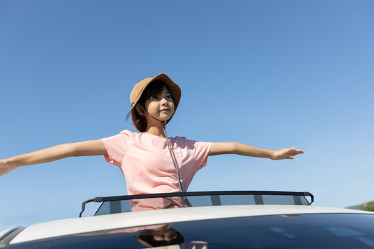 Asian girl with arms outstretched standing through a car sunroof during outdoor travel, showing childhood joy, freedom, and relaxed travel lifestyle.