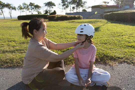 Asian mother removes a safety helmet from her young daughter while sitting outdoors after activity, showing parental care, child safety, and family bonding.