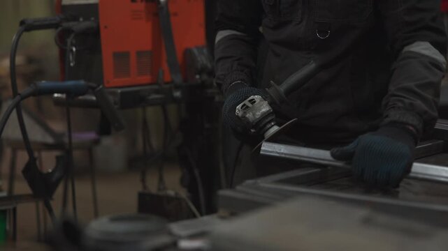Gloved workshop worker grinds metal bar with handheld tool, sending small orange sparks across dim industrial workspace, framed in tight close view with shallow depth and gritty technical mood