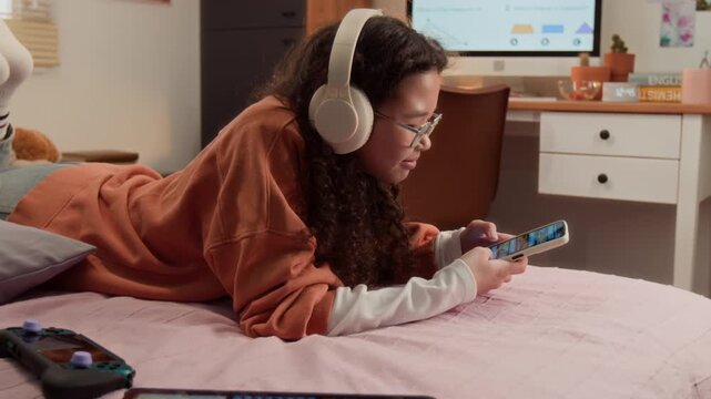East Asian teenager wearing headphones lying on bed and using smartphone. Tablet and controller nearby suggest gaming lifestyle, perfect for youth technology and entertainment marketing