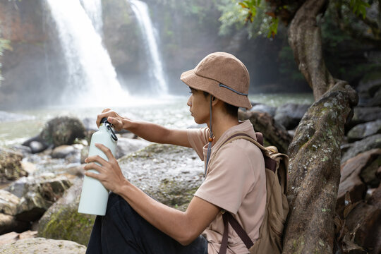 Medium Shot of Asian Teenage Boy Resting with Water Bottle Near Waterfall