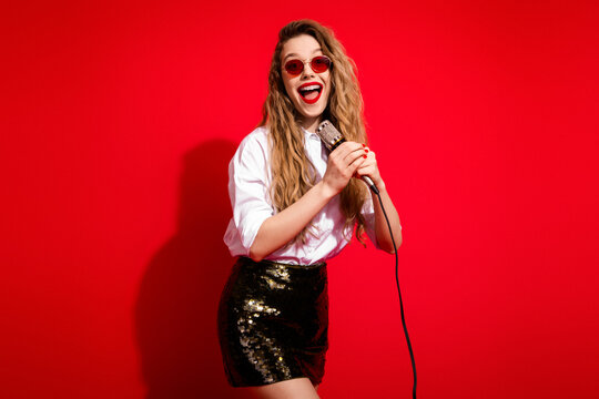 Young female singer in stylish attire with microphone performing joyfully against a vivid red background
