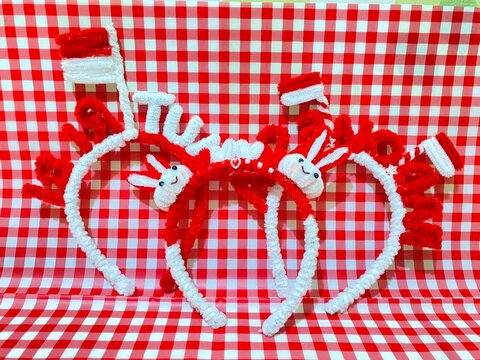 Handmade red and white pipe cleaner headbands with bow and flower decorations wrapped in clear plastic packaging, displayed against a red and white checkered pattern background.