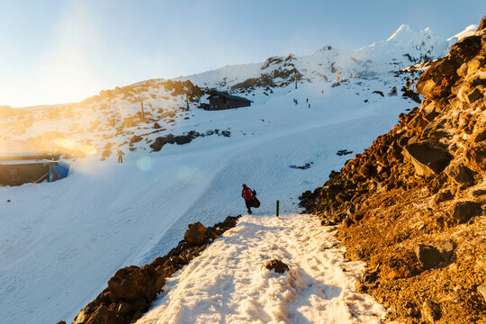 A hiker in a red jacket walks uphill through glowing sunrise light on the snowy Whakapapa slopes, with alpine huts scattered across the rugged terrain