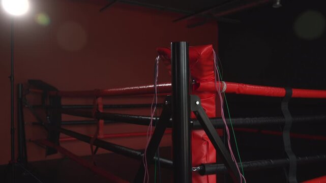 Empty boxing ring corner with red padded post, dark ropes, hanging cables, glossy metal support frame, and dramatic studio lighting in moody industrial sports setting