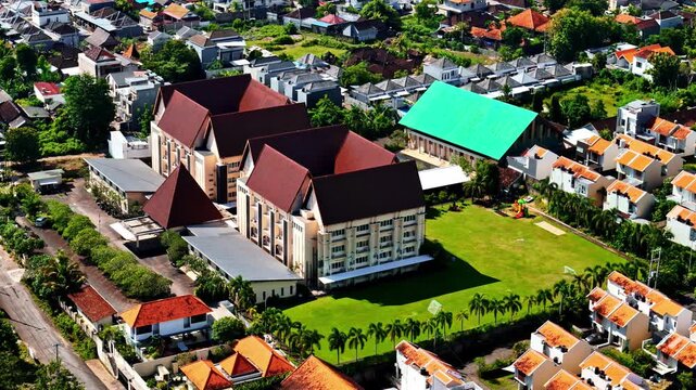 Aerial view of a large building complex with brown roofs and a green lawn surrounded by residential houses in Bali, Indonesia.