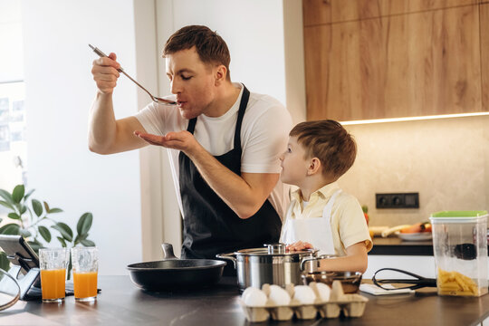 Caucasian male chef tastes food from a spoon while cooking in a modern kitchen with a young boy observing, various ingredients and utensils on the counter