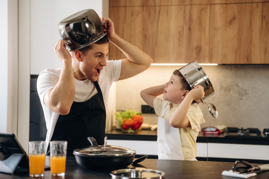 Father and son wearing stainless steel cooking pots on their heads playfully pose in a modern kitchen with fresh vegetables and orange juice on the counter