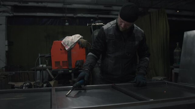 Skilled worker bends over steel panel during bright welding action inside dim industrial workshop with heavy machinery, glowing sparks, dark protective clothing, gritty metal surfaces, focused work