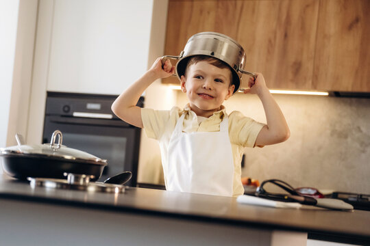 Young boy wearing a stainless steel pot on his head smiles while standing in a modern kitchen with cooking utensils and appliances visible in the background