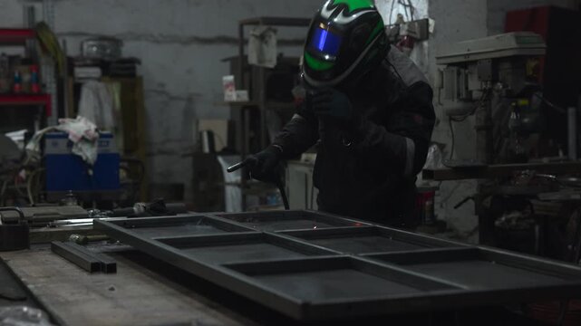 Worker in protective welding helmet and gloves shaping steel structure inside dim industrial workshop with blue arc glow, flying sparks, rough concrete walls, machinery, tools, and gritty