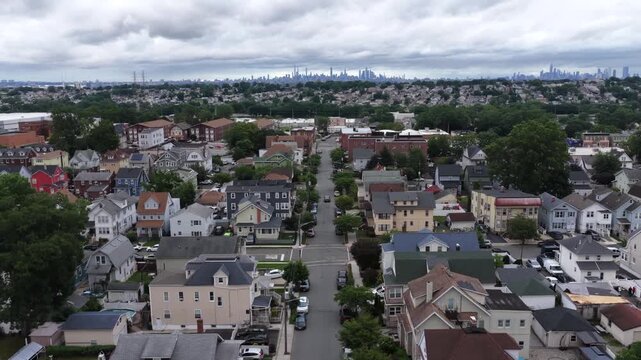 Dense Victorian-style houses line straight residential street in Belleville New Jersey, distant hazy Manhattan skyline rises beyond rooftops beneath cloudy overcast sky. Aerial drone rising wide shot