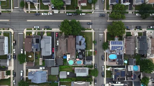 Backyard swimming pools cluster between dense rowhouses in Belleville New Jersey, parked cars line straight residential street beneath green trees and solar rooftops. Aerial drone top down
